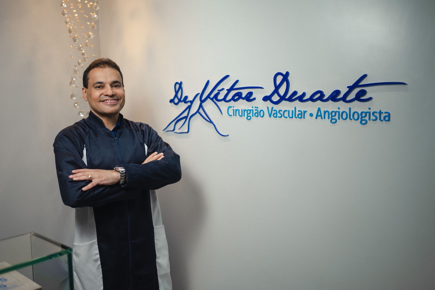 Dr. Vitor Duarte de jaleco em seu consultório, ao lado da sua marca — Cirurgião Vascular e Angiologista